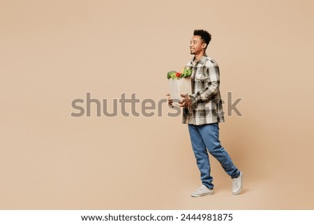 Full body side view profile young man wear grey shirt hold paper bag for takeaway mock up with food products go look aside isolated on plain beige background. Delivery service from shop or restaurant