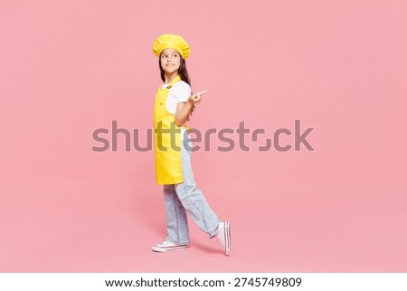 Full body side view small kid child girl housewife chef cook baker mother's helper wears yellow apron toque cap point index finger aside on area isolated on plain pink background. Cooking food concept