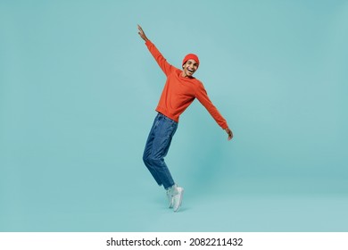 Full body side view smiling fun cheerful young happy african american man in orange shirt hat stand on toes leaning back dancing fooling around isolated on plain pastel light blue background studio.