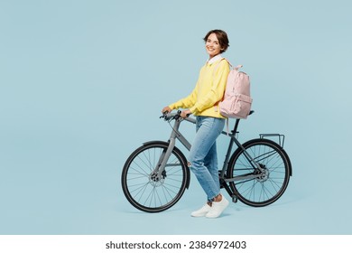 Full body side view smiling happy fun young woman student wear casual clothes sweater backpack bag ride bicycle look camera isolated on plain blue background. High school university college concept