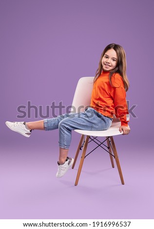 Full body side view of smiling little girl in orange sweatshirt and jeans with white sneakers sitting on chair in studio against violet background