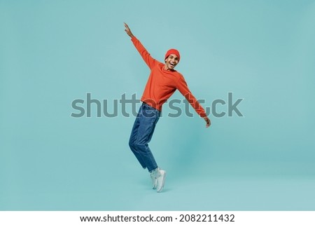 Full body side view smiling fun cheerful young happy african american man in orange shirt hat stand on toes leaning back dancing fooling around isolated on plain pastel light blue background studio.