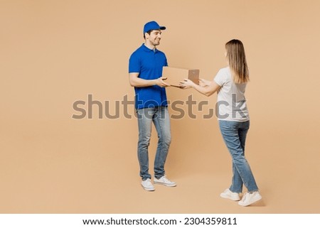 Full body side view smiling delivery guy employee man wear blue cap t-shirt uniform workwear work as dealer courier give woman cardboard box isolated on plain light beige background. Service concept