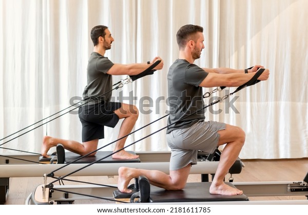 Full body side view of strong men in\
sportswear doing hug tree kneeling pilates exercise with resistance\
bands on reformer bed during training in\
gym