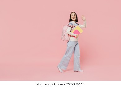 Full Body Side View Teen Fun Student Girl Of Asian Ethnicity Wearing Sweater Hold Backpack Books Walk Going Waving Hand Isolated On Pastel Plain Pink Background Education In University College Concept