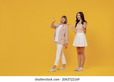 Full body side view two young amazed smiling happy daughter mother together couple women in casual beige clothes walk point index finger aside on workspace isolated on plain yellow background studio.