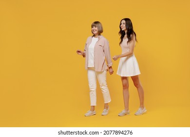 Full body side view two young smiling happy daughter mother together couple women in casual beige clothes looking camera walk isolated on plain yellow color background studio. Family lifestyle concept