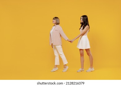 Full Body Side View Two Young Smiling Happy Daughter Mother Together Couple Women Wear Casual Beige Clothes Hold Hands Walk Like Follow Me Isolated On Plain Yellow Color Background Studio Portrait.