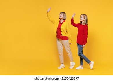 Full Body Side View Woman 50s In Red Shirt Have Fun With Teenager Girl 12-13 Years Old. Grandmother Granddaughter Walk Going Waving Hand Isolated On Plain Yellow Background. Family Lifestyle Concept.