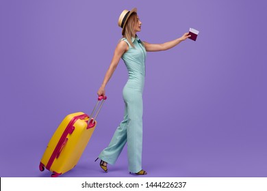 Full body side view of young woman with passport and ticket pulling suitcase and walking against purple background having flight