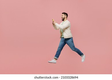 Full Body Side View Young Happy Man 20s Wearing Trendy Jacket Shirt Hold In Hand Use Mobile Cell Phone Jump High Walk Go Isolated On Plain Pastel Light Pink Background Studio. People Lifestyle Concept