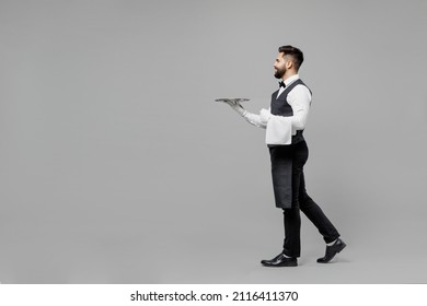 Full body side view young barista male waiter butler man in white shirt vest elegant uniform work at cafe hold in hand carrying metal tray isolated on plain grey background Restaurant employee concept