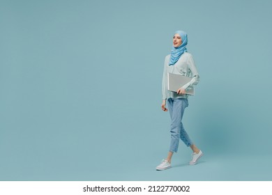 Full body side view young arabian asian muslim woman in abaya hijab hold closed laptop pc computer walking isolated on plain blue background studio. People uae middle eastern islam religious concept.