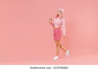 Full body side view young woman 20s with bright dyed rose hair in rosy top shirt hat hold takeaway delivery craft paper brown cup coffee to go walk going isolated on plain light pastel pink background