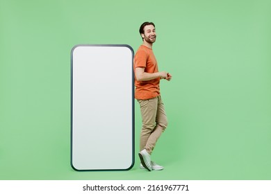 Full body side view young man wear casual orange t-shirt stand near mobile cell phone with blank screen workspace area isolated on plain pastel light green color background. People lifestyle concept