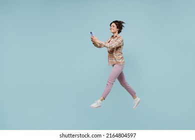 Full Body Side View Young Smiling Happy Woman 20s Wearing Casual Brown Shirt Hold In Hand Use Mobile Cell Phone Jump High Isolated On Pastel Plain Light Blue Color Background People Lifestyle Concept