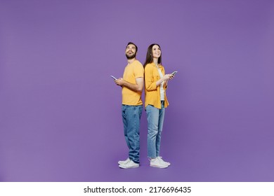 Full body side view young couple two friends family man woman together in yellow clothes hold in hand use mobile cell phone stand back to back browsing internet isolated on plain violet background. - Shutterstock ID 2176696435
