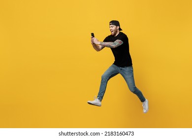 Full body side view young bearded tattooed man 20s he wearing casual black t-shirt cap jump high run fast hold in hand use mobile cell phone isolated on plain yellow wall background studio portrait.