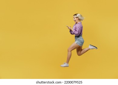 Full Body Side View Young Blonde Woman 20s Wear Pink Tied Shirt White T-shirt Jump High Hold In Hand Use Mobile Cell Phone Run Fast Isolated On Plain Yellow Background Studio. People Lifestyle Concept