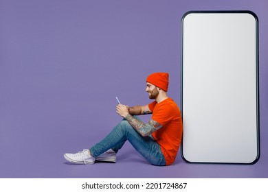 Full body side view young man in red hat t-shirt use smartphone sit near big huge blank screen mobile cell phone workspace mockup isolated on plain pastel purple background People lifestyle concept - Shutterstock ID 2201724867