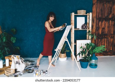 Full Body Side View Of Young Female Artist Standing Near Easel With Paintbrush And Colors While Looking At Painting On Canvas In Creative Studio