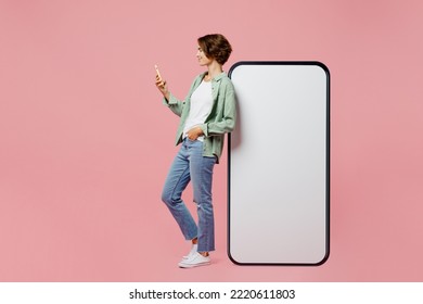 Full body side view young woman wear green shirt white t-shirt near big huge blank screen mobile cell phone smartphone with mockup use mobile cell phone isolated on plain pastel light pink background - Shutterstock ID 2220611803