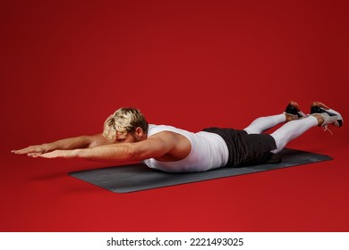 Full Body Side View Young Strong Sporty Toned Sportsman Man Wear White Clothes Spend Time In Home Gym Lie Face Down Raise Up Hands Legs Isolated On Plain Red Background. Workout Sport Fit Body Concept