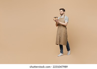 Full body side view young man barista barman employee in brown apron work in shop hold craft paper brown cup coffee to go walk isolated on plain pastel beige background Small business startup concept - Shutterstock ID 2231047989