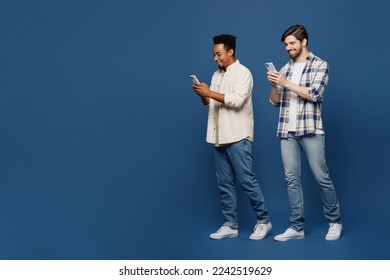Full body side view young two friends happy smiling cheerful men 20s wear white casual shirts together hold in hand use mobile cell phone walk go stroll isolated plain dark royal navy blue background