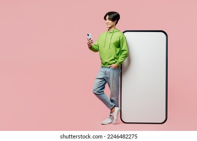 Full body side view young man of Asian ethnicity wear green hoody big huge blank screen mobile cell phone with area hold in hand use smartphone isolated on plain pastel light pink background studio - Shutterstock ID 2246482275