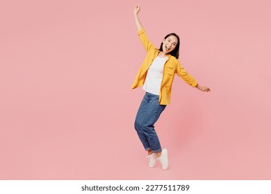 Full body side view young woman of Asian ethnicity wears yellow shirt white t-shirt leaning back with outstretched hands stand on toes isolated on plain pastel light pink background studio portrait