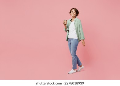 Full body side view young woman 20s she wear green shirt white t-shirt hold takeaway delivery craft paper brown cup coffee to go isolated on plain pastel light pink background People lifestyle concept