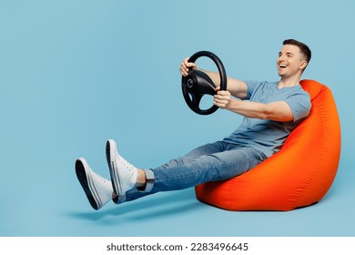 Full body side view young happy man wear casual t-shirt sit in bag chair hold steering wheel pretend driving car isolated on plain pastel light blue cyan background studio portrait. Lifestyle concept