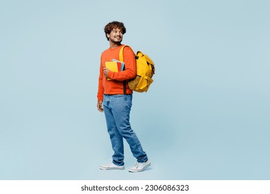 Full body side view young teen Indian boy student he wearing casual clothes backpack bag hold books walk go looka aside isolated on plain pastel blue background. High school university college concept