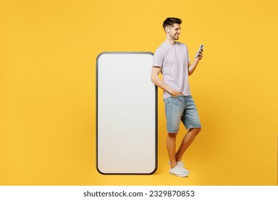 Full body side view young smiling man he wears light purple t-shirt casual clothes big huge blank screen mobile cell phone with area using smartphone chat online isolated on plain yellow background - Powered by Shutterstock