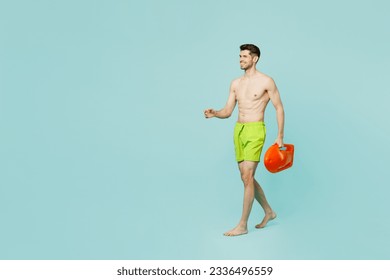 Full body side view young lifeguard man wear green shorts swimsuit relax near hotel pool hold lifebuoy walk go look aside isolated on plain blue background. Summer vacation sea rest sun tan concept - Powered by Shutterstock