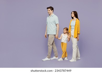 Full body side view young parents mom dad child kid daughter girl 6 years old wearing blue yellow casual clothes hold hands walk go look aside isolated on plain purple background. Family day concept