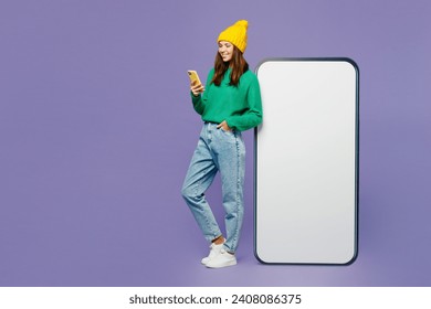 Full body side view young woman she wears green sweater yellow hat casual clothes big huge blank screen mobile cell phone with area using smartphone isolated on plain pastel light purple background - Powered by Shutterstock