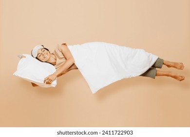 Full body side view young Latin woman wears pyjamas jam sleep eye mask rest relax at home fly fall hover over air on pillow under duvet isolated on plain beige background. Good mood night nap concept - Powered by Shutterstock