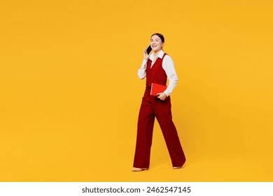 Full body side view young lawyer employee business woman of Asian ethnicity wearing red vest shirt work at office talk speak on mobile cell phone walk go hold book isolated on plain yellow background - Powered by Shutterstock