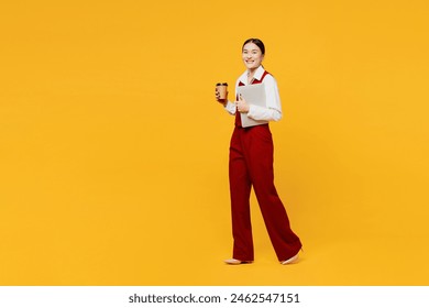 Full body side view young lawyer employee business woman of Asian ethnicity wear formal red vest shirt work at office hold closed laptop pc computer drink coffee isolated on plain yellow background - Powered by Shutterstock