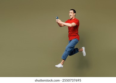 Full body side view young middle eastern man wear red t-shirt casual clothes jump high hold in hand use mobile cell phone isolated on plain pastel green background studio portrait. Lifestyle concept