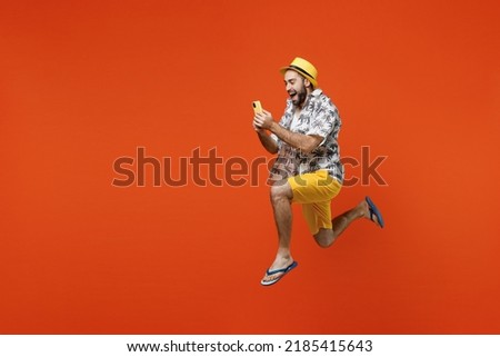 Full body side view young tourist man wear beach shirt hat jump high run fast use hold mobile cell phone isolated on plain orange background studio portrait. Summer vacation sea rest sun tan concept
