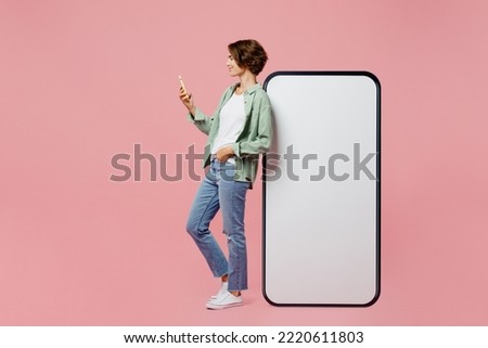 Full body side view young woman wear green shirt white t-shirt near big huge blank screen mobile cell phone smartphone with mockup use mobile cell phone isolated on plain pastel light pink background