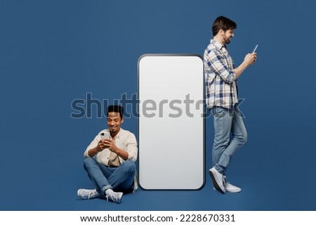 Full body side view young two friend men wear casual shirts together sit near big huge blank screen mobile cell phone gadget with mockup use smartphone isolated plain dark royal navy blue background