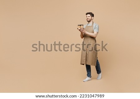 Full body side view young man barista barman employee in brown apron work in shop hold craft paper brown cup coffee to go walk isolated on plain pastel beige background Small business startup concept