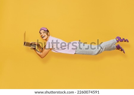 Full body side view young woman wear purple pyjamas jam sleep eye mask rest relax at home fly fall hover over air hurry up use laptop pc computer isolated on plain yellow background Night nap concept