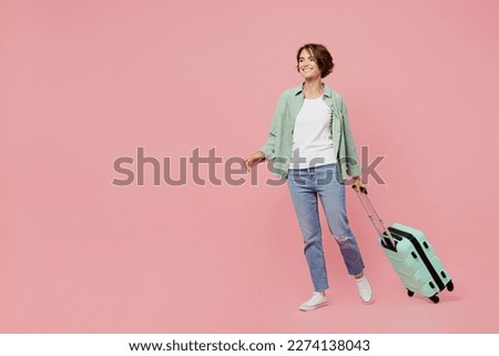 Full body side view young traveler woman wear green shirt hold suitcase isolated on plain pastel light pink background Tourist travel abroad in free time rest getaway Air flight trip journey concept