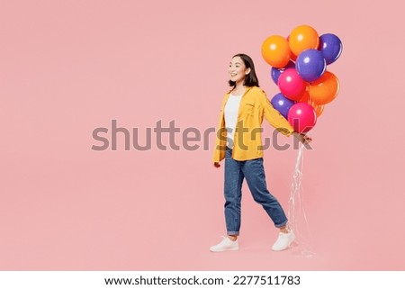 Full body side view young woman of Asian ethnicity wear yellow shirt white t-shirt hold bunch of colorful air balloons walk go isolated on plain pastel light pink background studio. Lifestyle concept