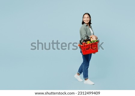 Full body side view young woman wear casual clothes hold red basket with food products look aside on area isolated on plain blue background studio portrait. Delivery service from shop or restaurant